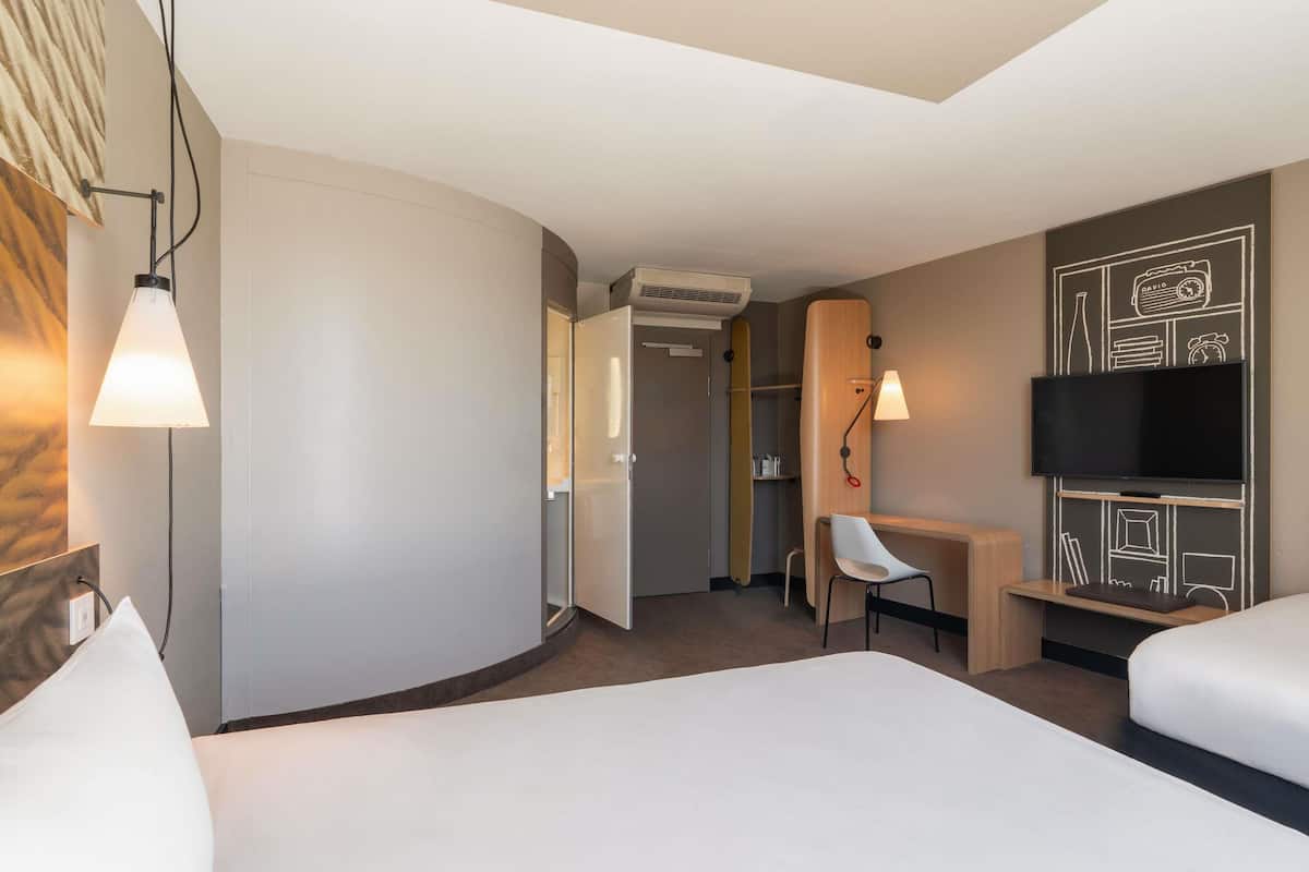 A hotel room with a bed, a desk, a chair, a TV, and an air conditioning unit.