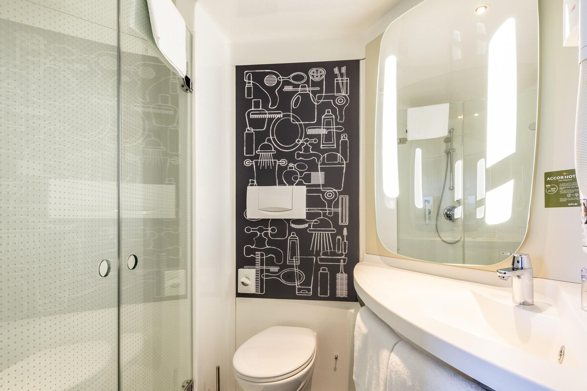 Une salle de bain moderne avec une douche à parois de verre, des toilettes blanches et un grand miroir au-dessus d’un lavabo blanc.