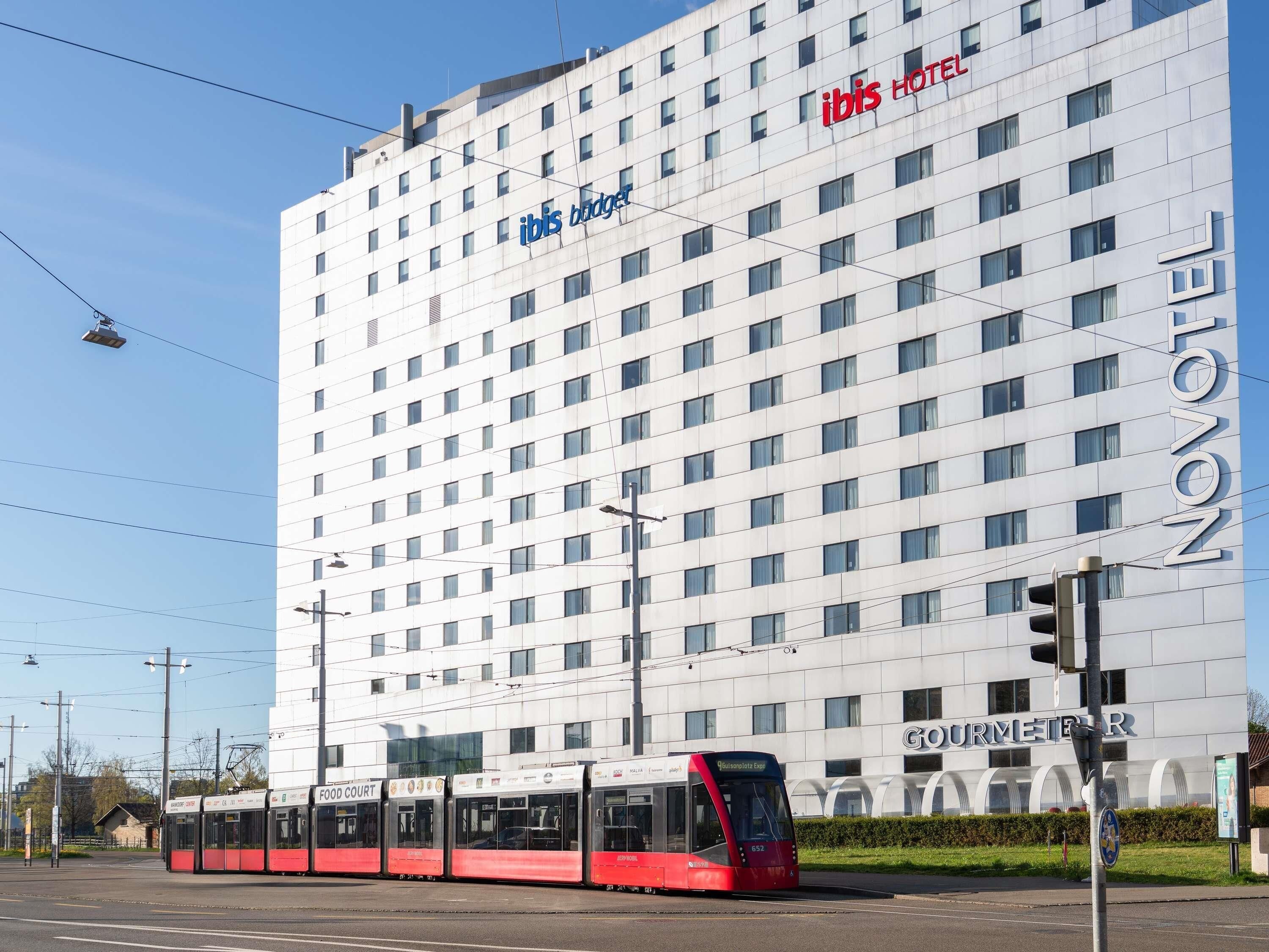 Un bâtiment d’hôtel moderne, avec une enseigne bien visible indiquant « hôtel ibis », une ligne de tramway et une enseigne indiquant un « espace restauration ».