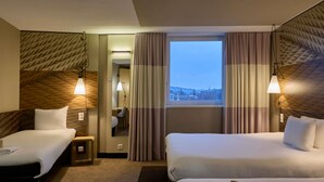 Een hotelkamer met twee bedden, een bureau, een stoel, een raam met gordijnen en uitzicht op de stad.