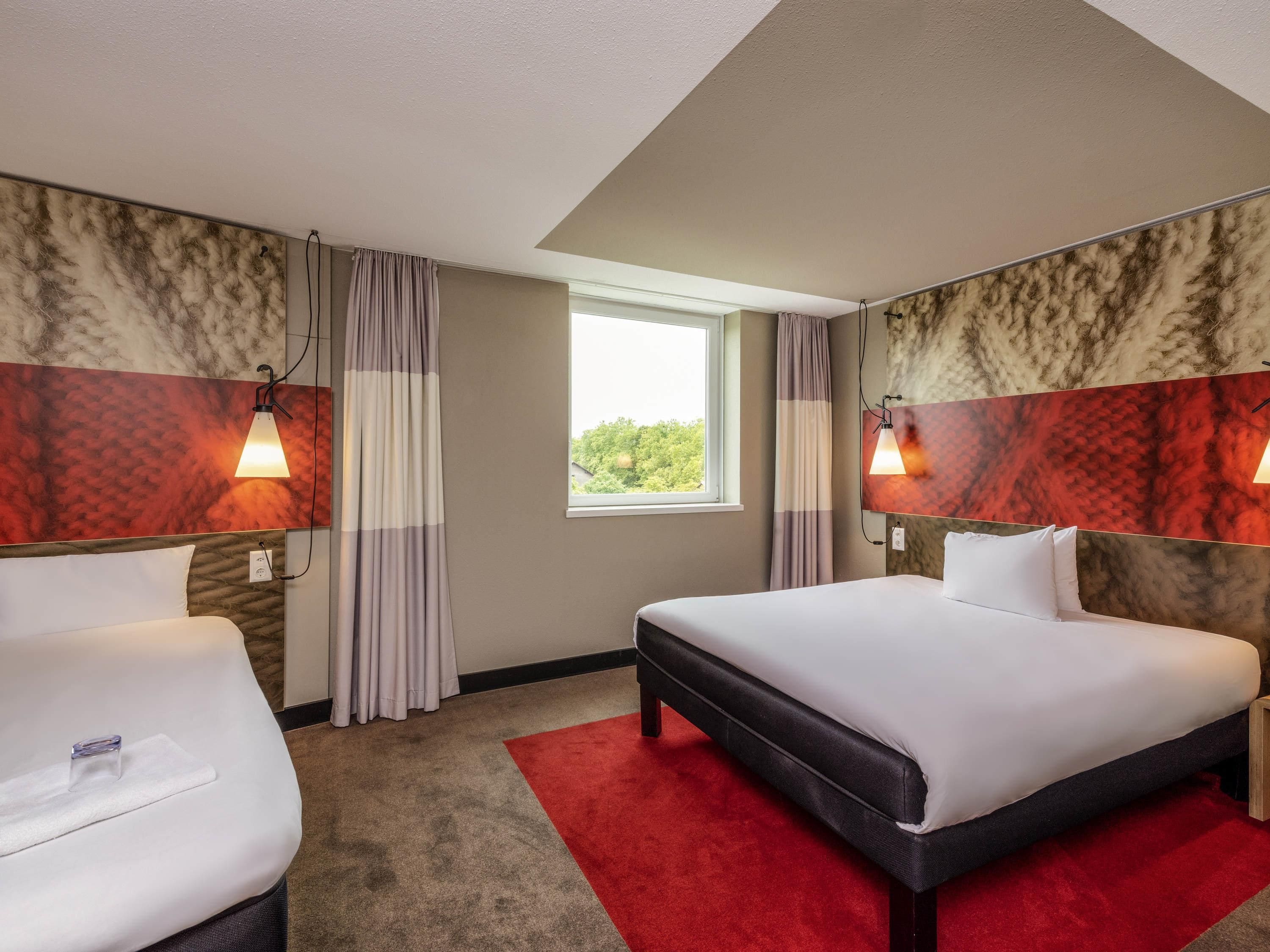 A hotel room with two beds, a red carpet, and a window with a view of greenery.