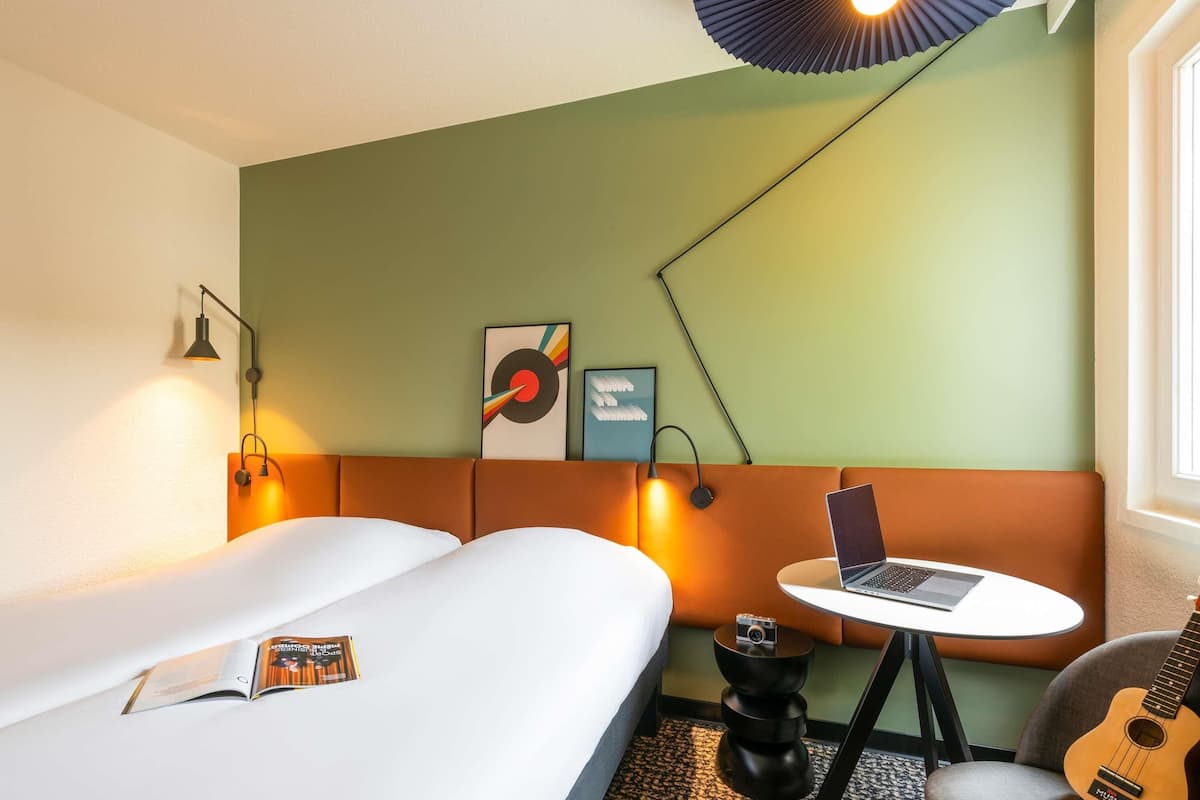 Une chambre d’hôtel avec un lit, une petite table sur laquelle se trouve un ordinateur portable, une guitare et des œuvres d’art accrochées au mur.