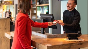 A modern bar setting with a wooden counter, a digital menu board, and a cash register. - ibis Nuits Saint Georges (Nuits-Saint-Georges)