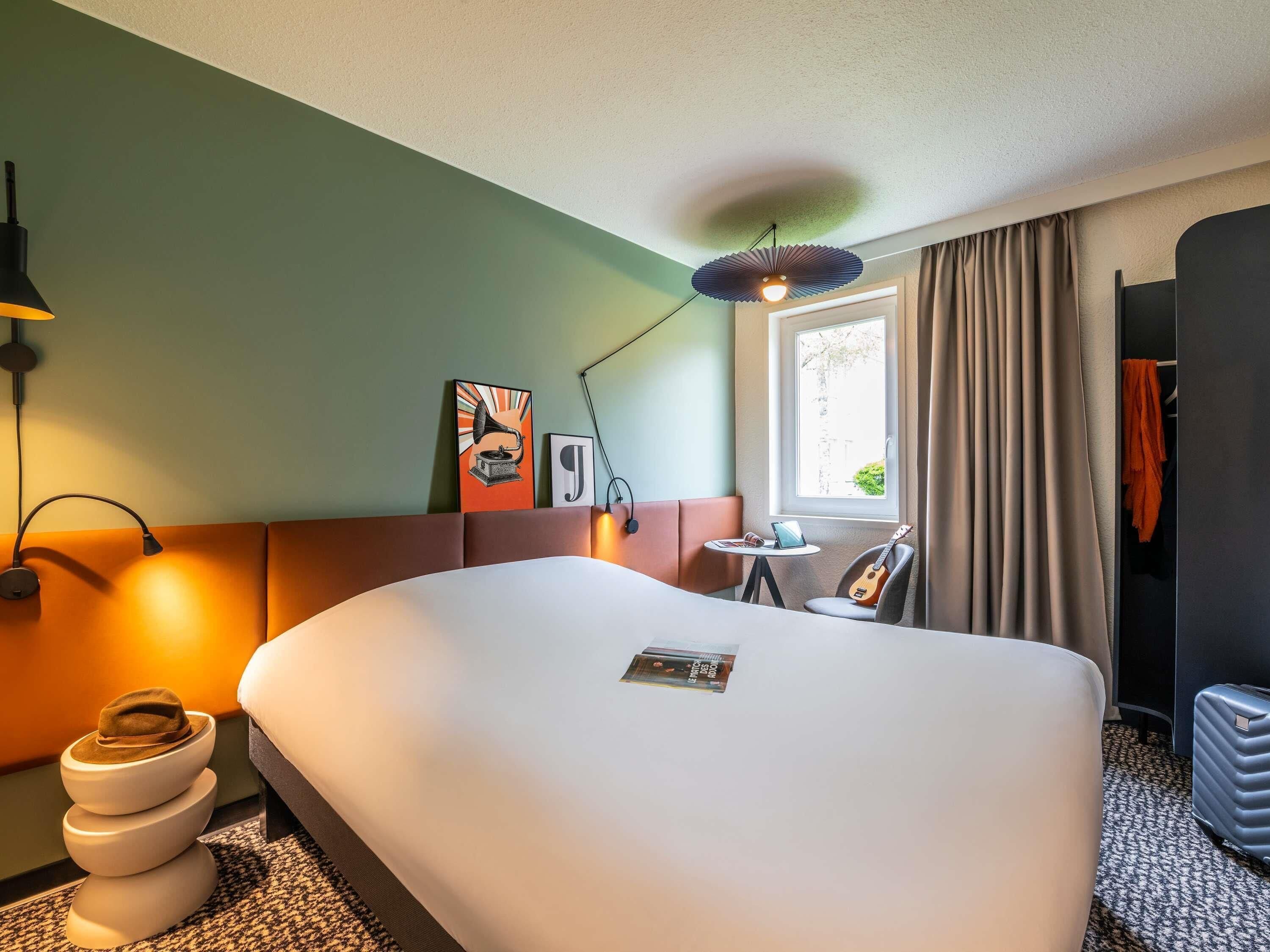 A hotel room with a large bed, a bedside table with a lamp, a window with curtains, and a view of the outside.