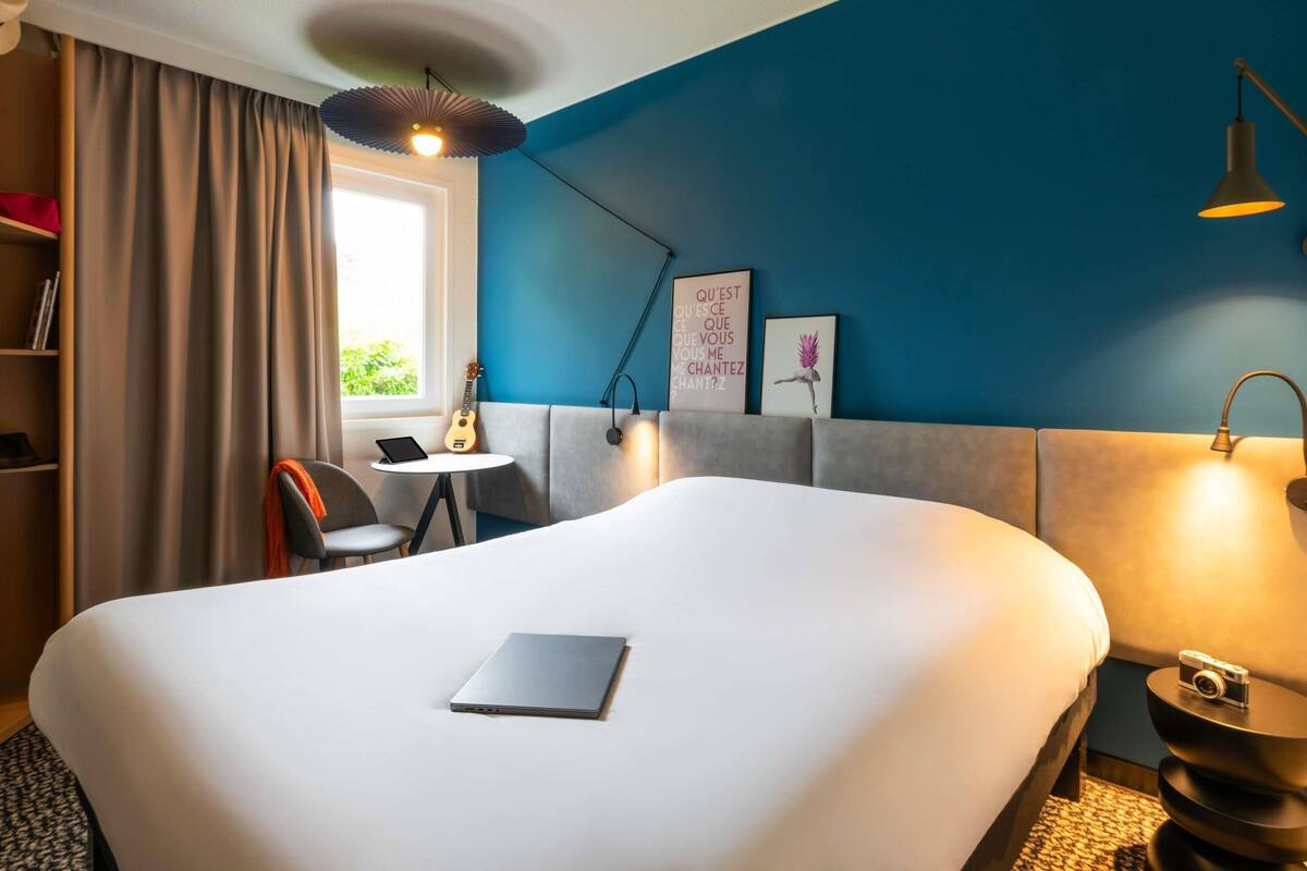Une chambre d’hôtel avec un grand lit, un bureau avec un ordinateur portable, une guitare et une fenêtre avec des rideaux.