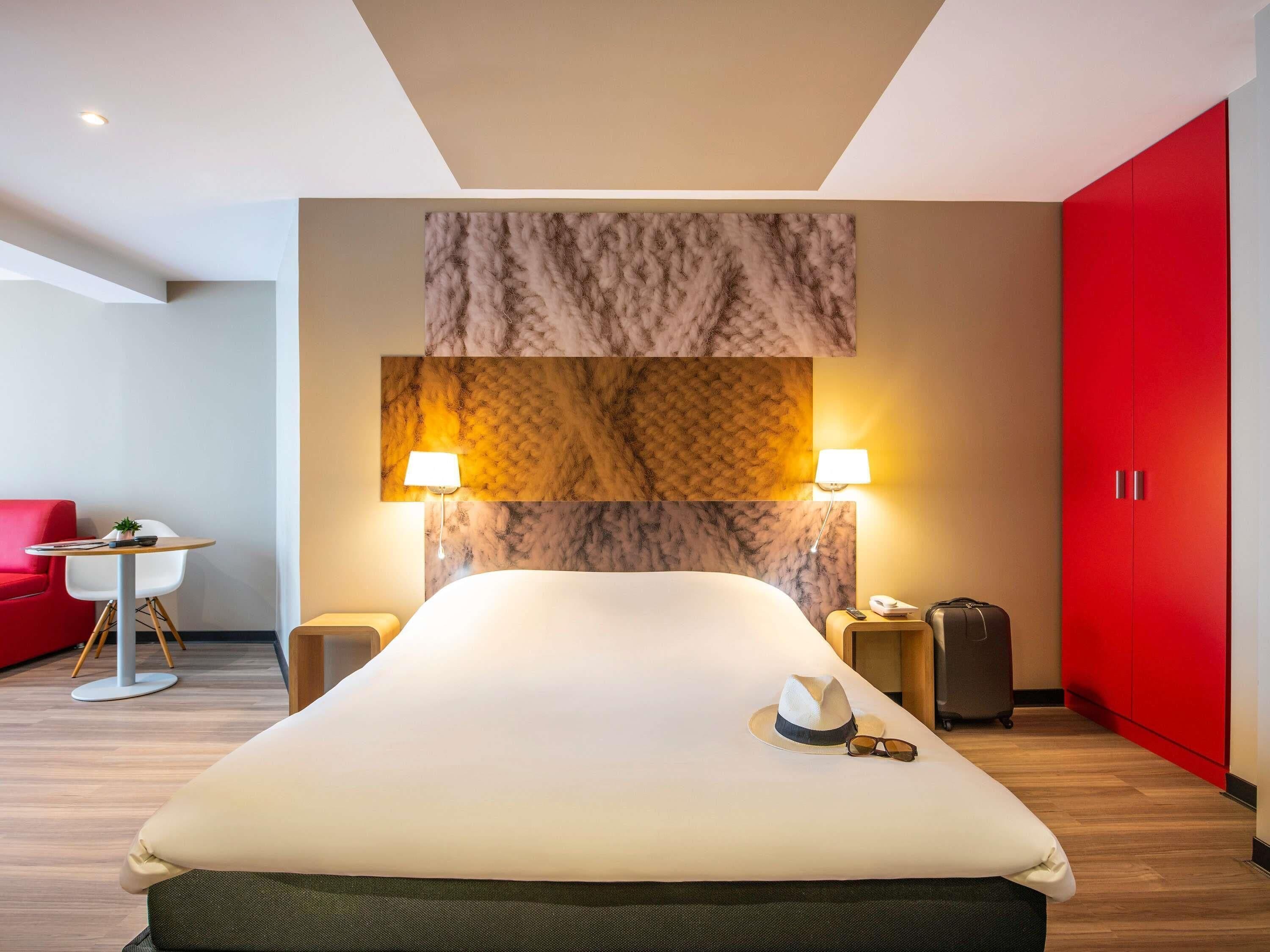 Habitación de hotel moderna con una cama grande, dos lámparas de noche, un armario rojo, una mesita con una silla y un sofá rojo.