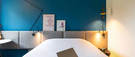 Una habitación de hotel moderna con una cama, una computadora portátil, una lámpara de pared, una cita enmarcada y una fotografía de una bailarina.