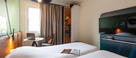Habitación de hotel con una cama grande, un escritorio con una laptop, una silla, una guitarra, una estantería y un televisor.
