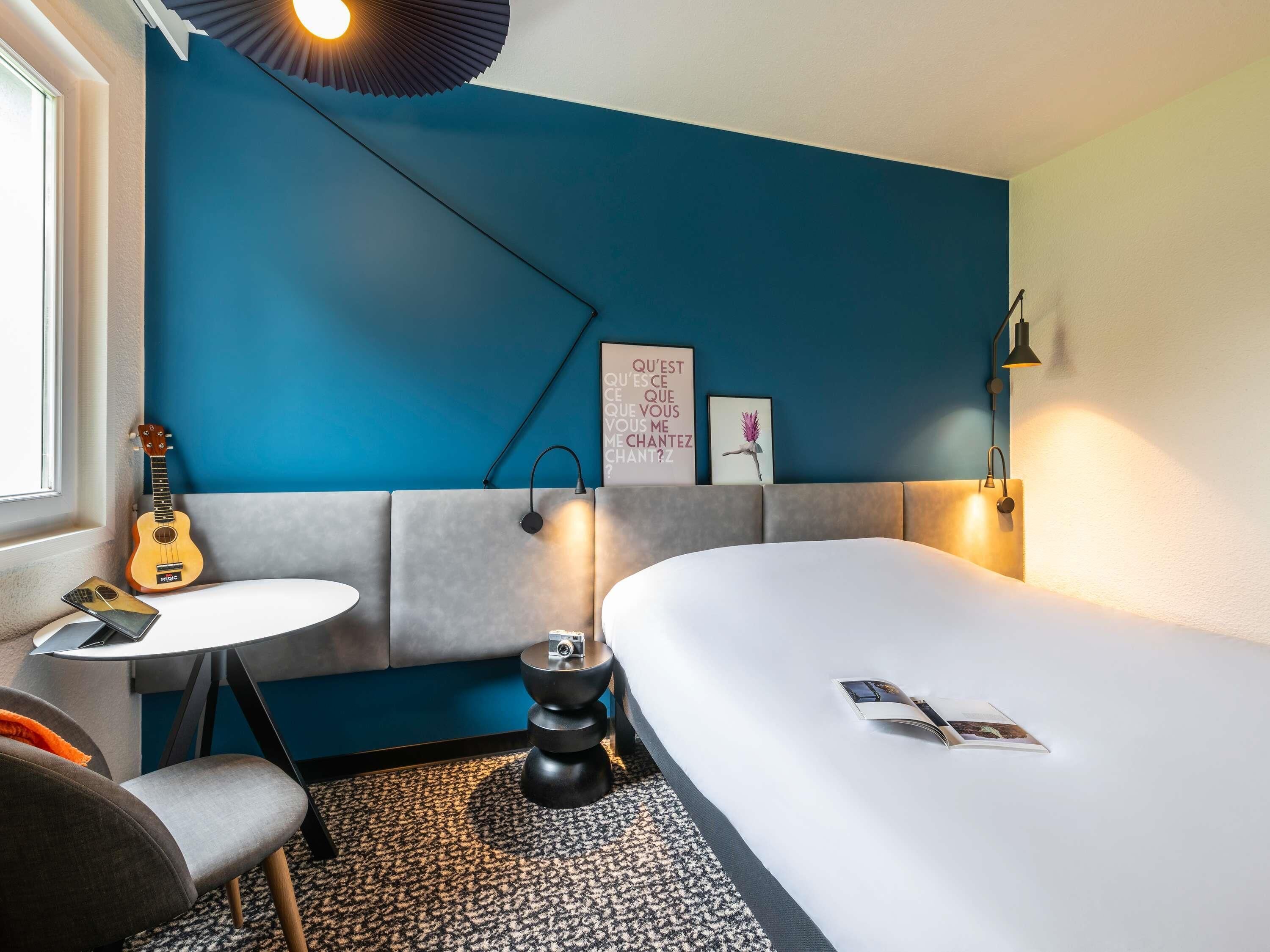 A modern hotel room with a blue accent wall, a bed, a small table with a guitar, and a wall-mounted lamp.