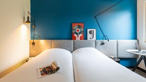 A modern room with a blue accent wall, a grey sofa, a small round table with a laptop, and a floor lamp. - ibis Nuits Saint Georges (Nuits-Saint-Georges)