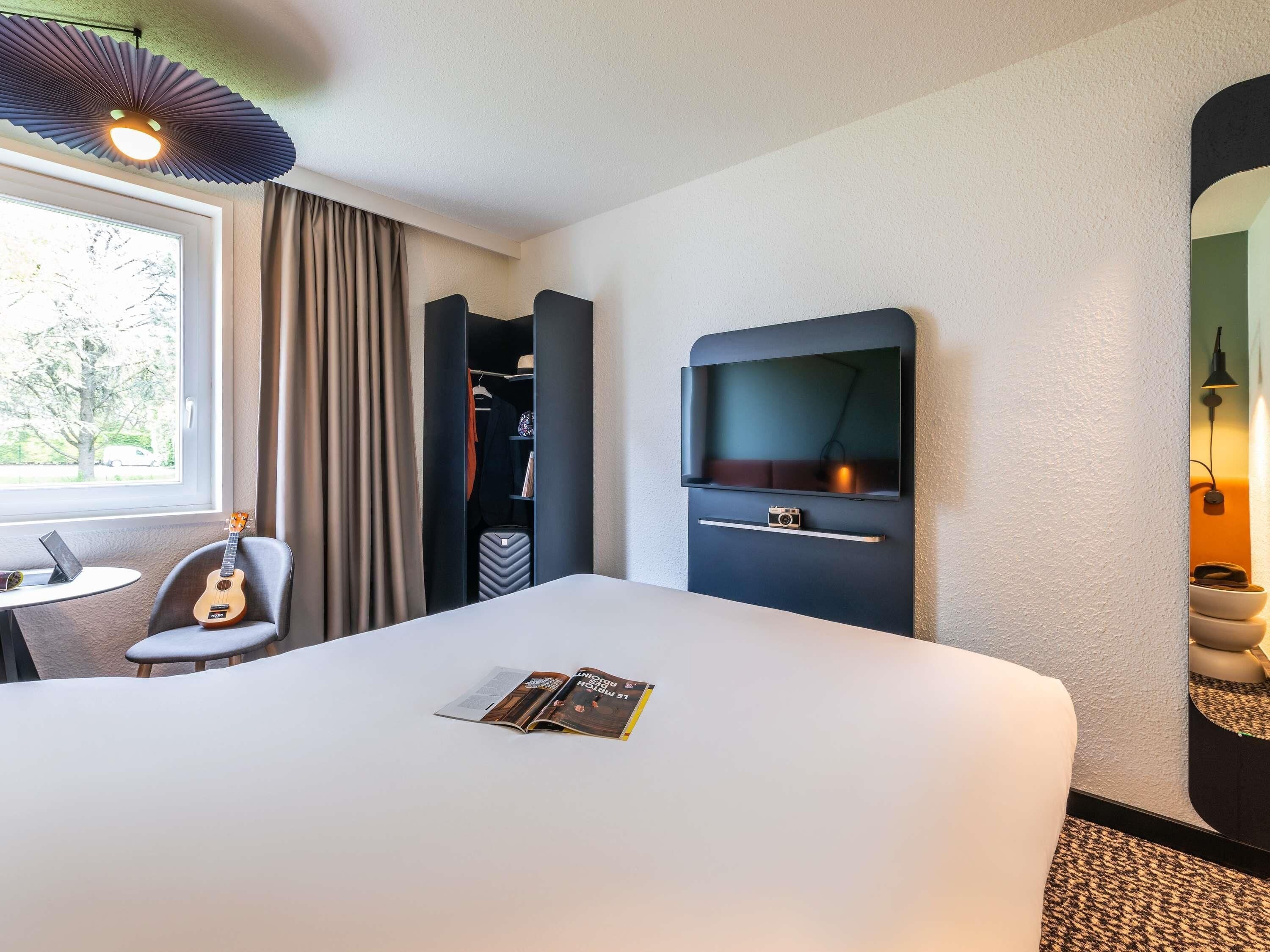 Een hotelkamer met een bed, een bureau met een laptop, een gitaar, een kledingkast, een televisie en een tijdschrift op het bed.