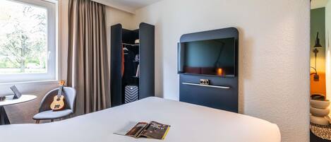 Een hotelkamer met een bed, een bureau met een laptop, een gitaar, een kledingkast, een televisie en een tijdschrift op het bed.