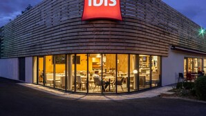 A modern building with a curved wooden facade and a prominent red 'ibis' sign. Inside, there are multiple seating areas with tables and chairs. - ibis Nuits Saint Georges (Nuits-Saint-Georges)
