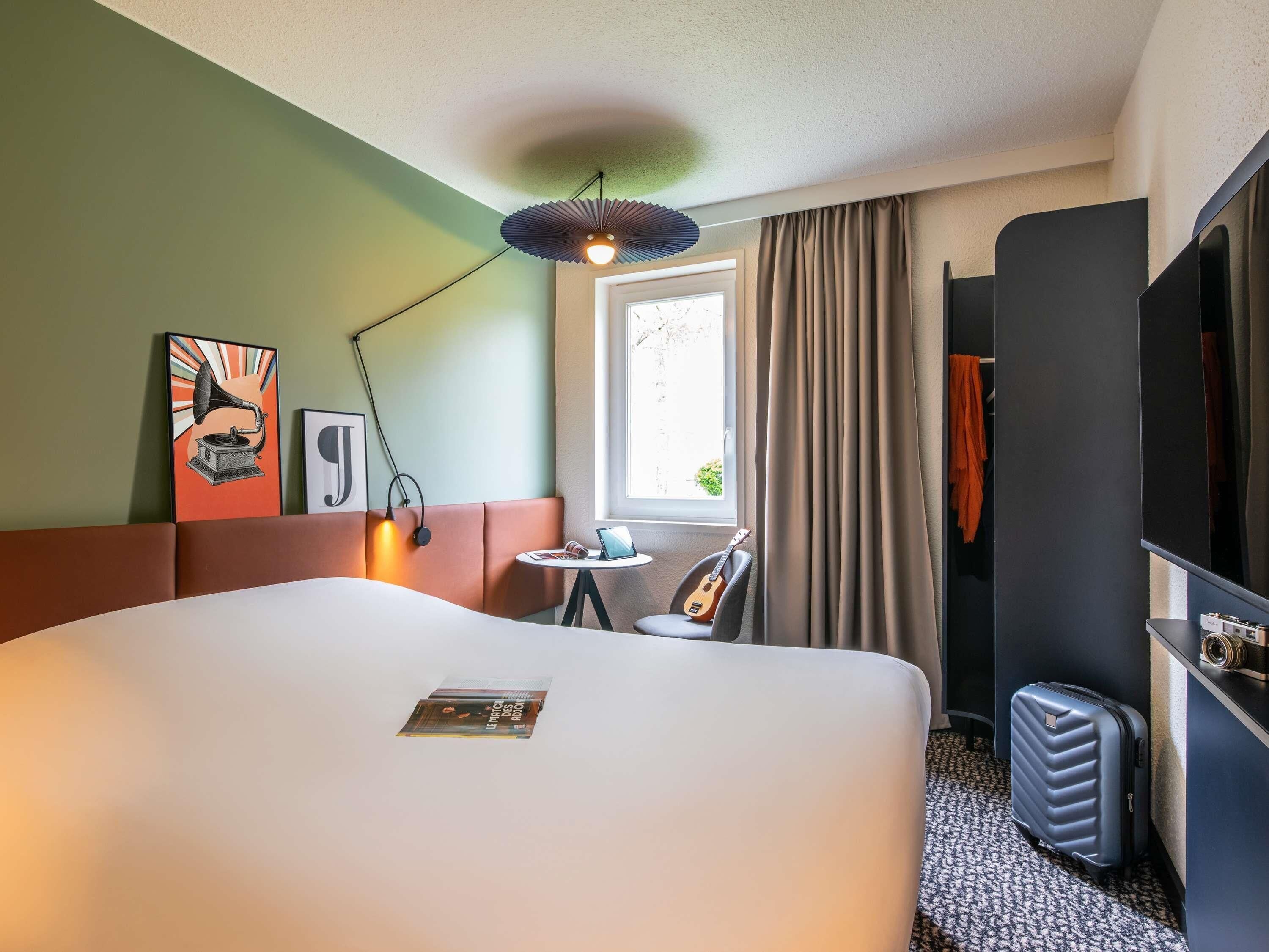 Une chambre d’hôtel avec un grand lit, un bureau avec un ordinateur portable, une guitare, une valise et une fenêtre avec des rideaux.