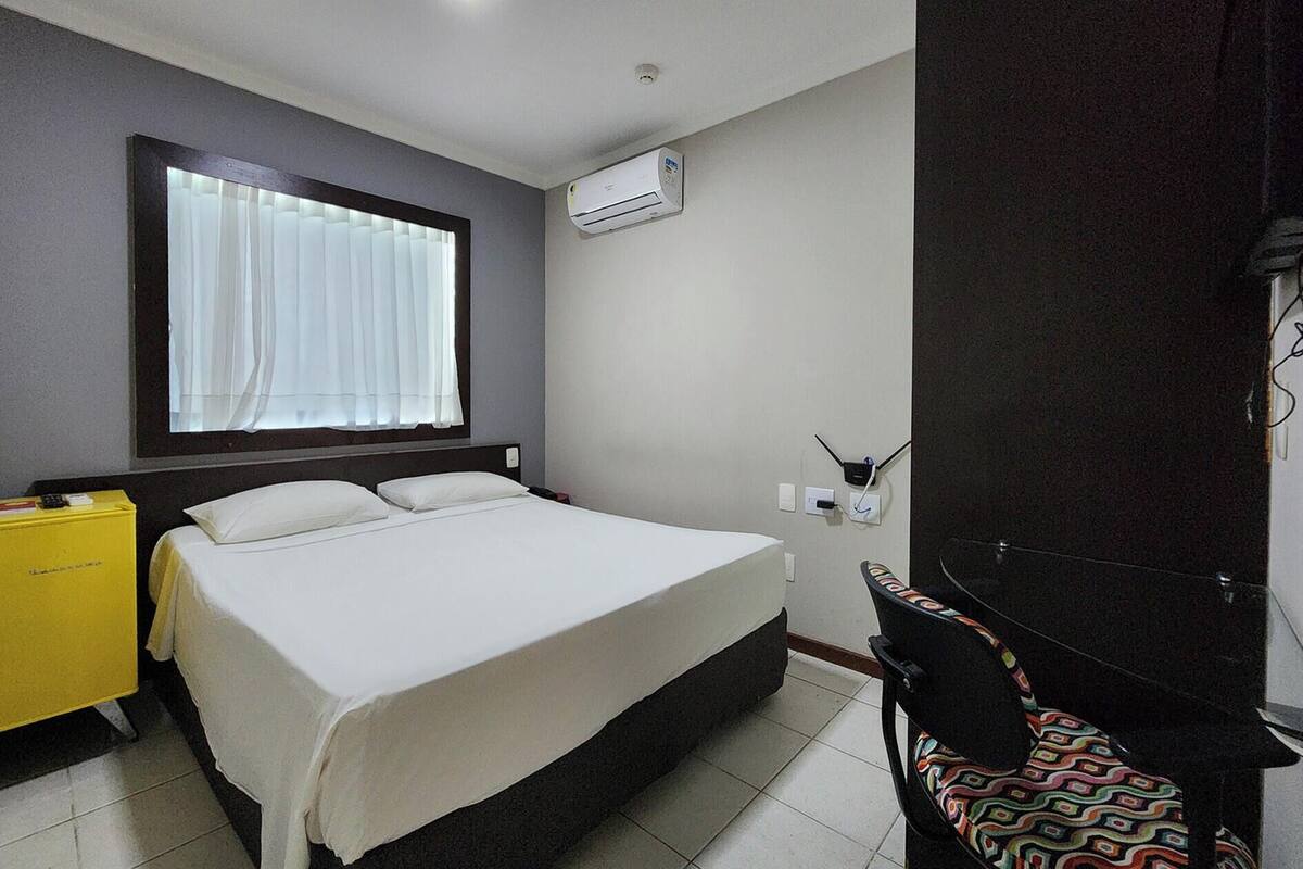 A hotel room with a bed, a window with curtains, an air conditioning unit, a desk with a chair, and a small table with a yellow box.