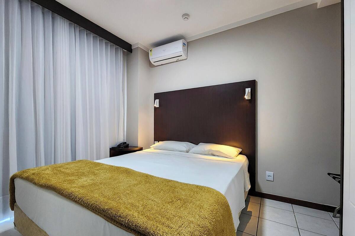 A hotel room with a bed, a nightstand, an air conditioning unit, and a window with curtains.