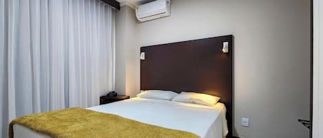 A hotel room with a bed, a nightstand, an air conditioning unit, and a window with curtains.