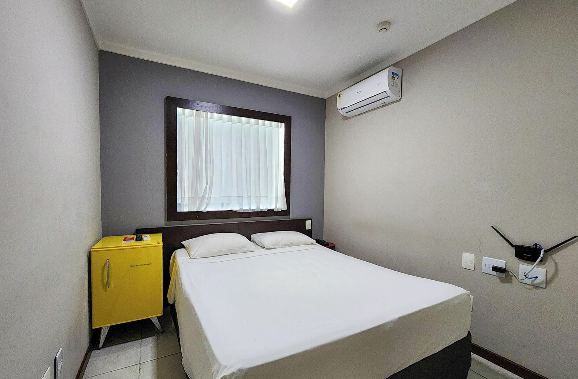A small, clean room with a bed, a yellow minibar, a window with white curtains, and an air conditioning unit.