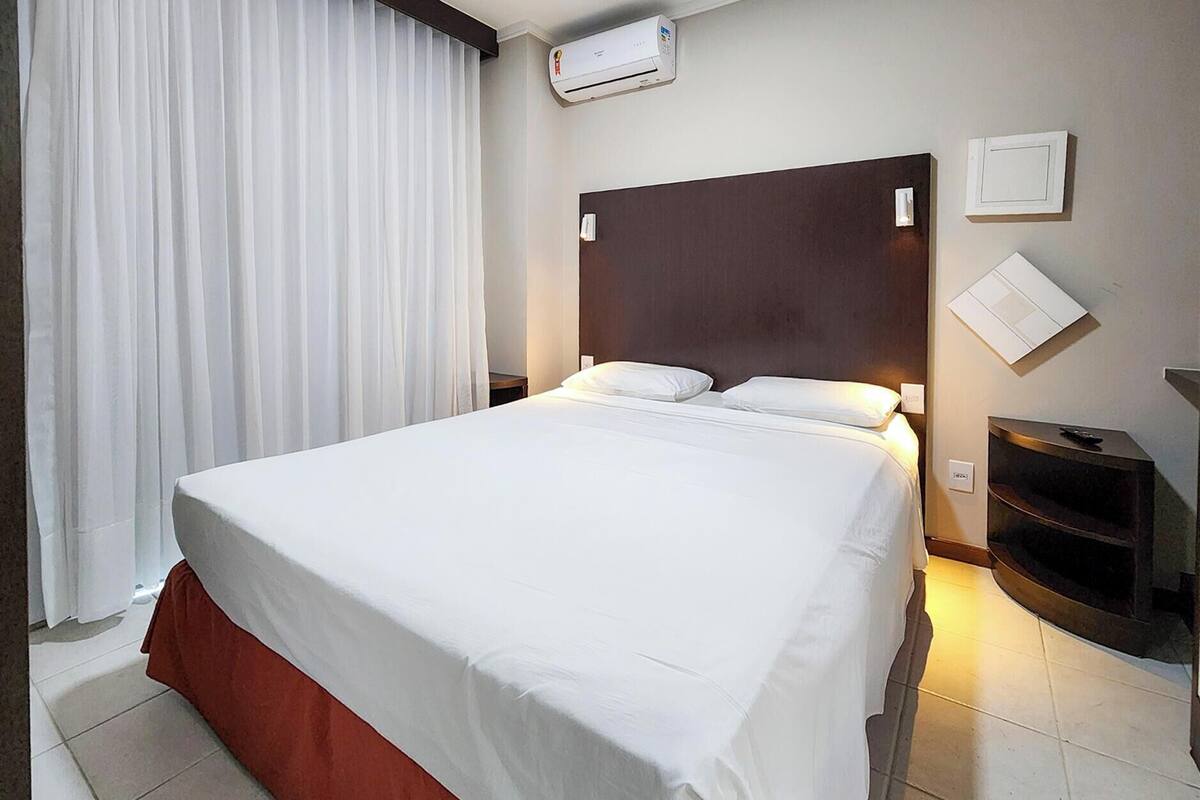 A hotel room with a large bed, white curtains, an air conditioning unit, a wooden headboard, a small table, and a TV.