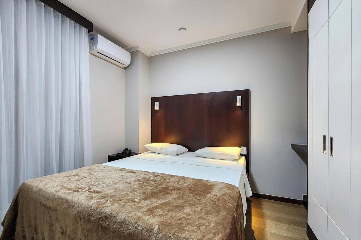 A hotel room with a bed, a wooden headboard, a nightstand, a telephone, and an air conditioning unit.