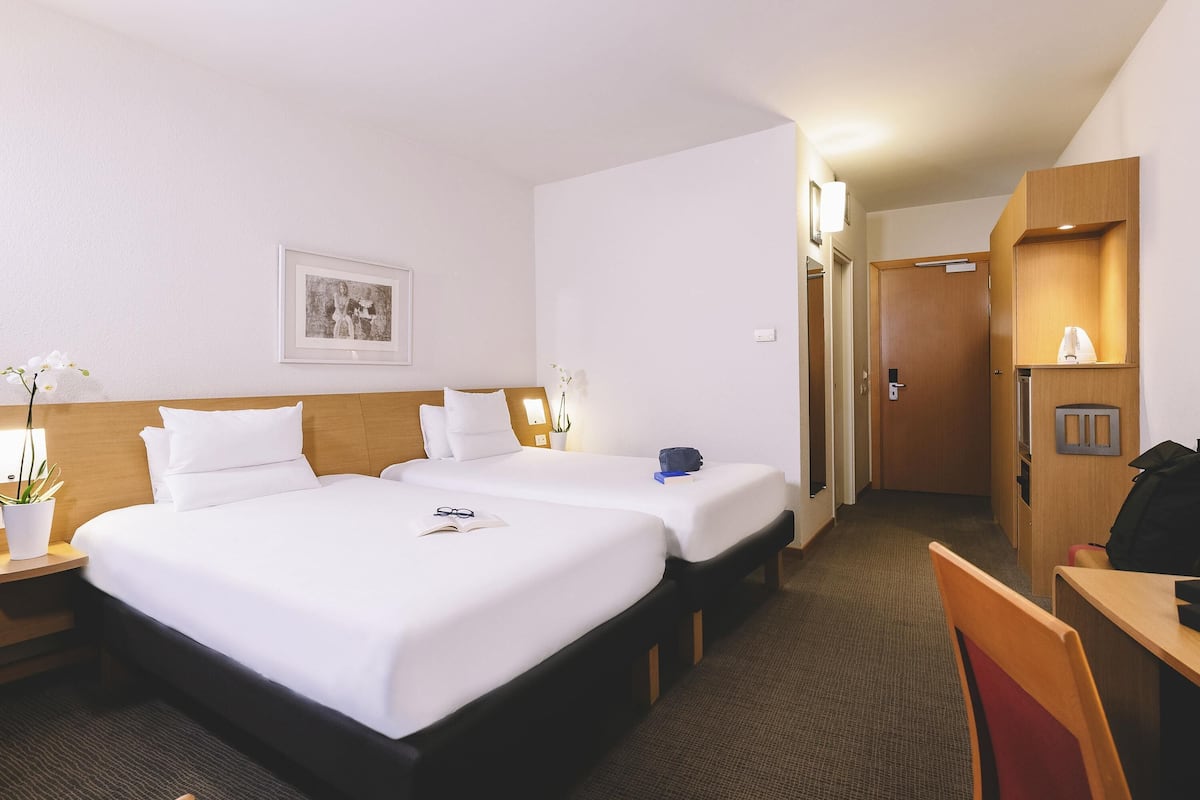 A hotel room with two beds, a desk, a chair, a small table, a potted plant, and a framed picture on the wall.