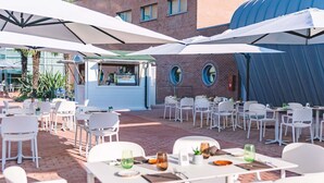 Un área de comedor al aire libre con mesas y sillas blancas, cubierta por grandes sombrillas, y un edificio de ladrillo al fondo.
