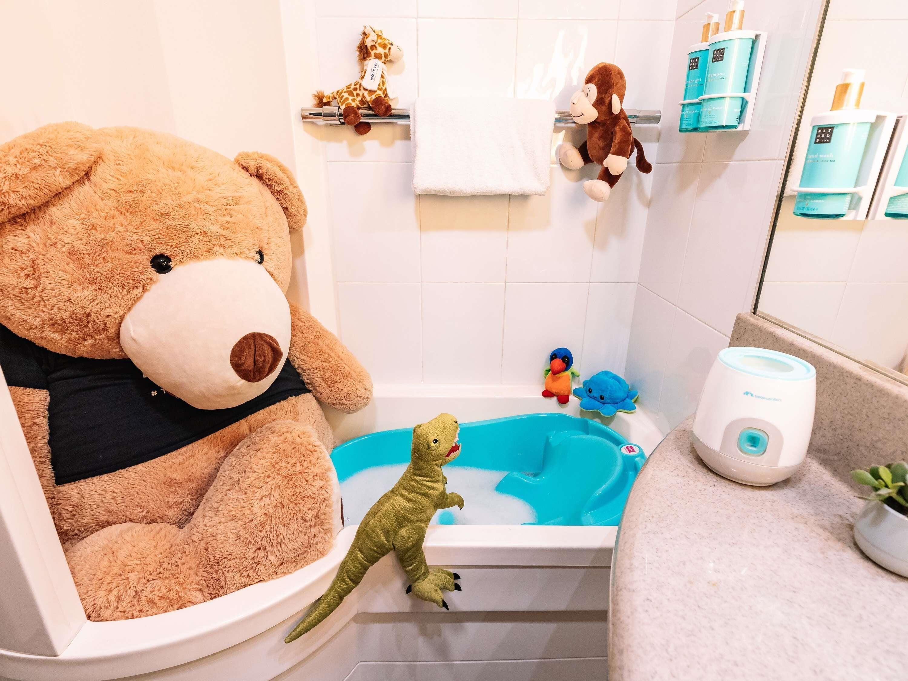 Une salle de bain avec une baignoire, un grand ours en peluche, un petit jouet de dinosaure et un petit singe en peluche.