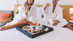 Dos personas vestidas de blanco sentadas en una cama con artículos de desayuno, como pasteles, frutas y bebidas.