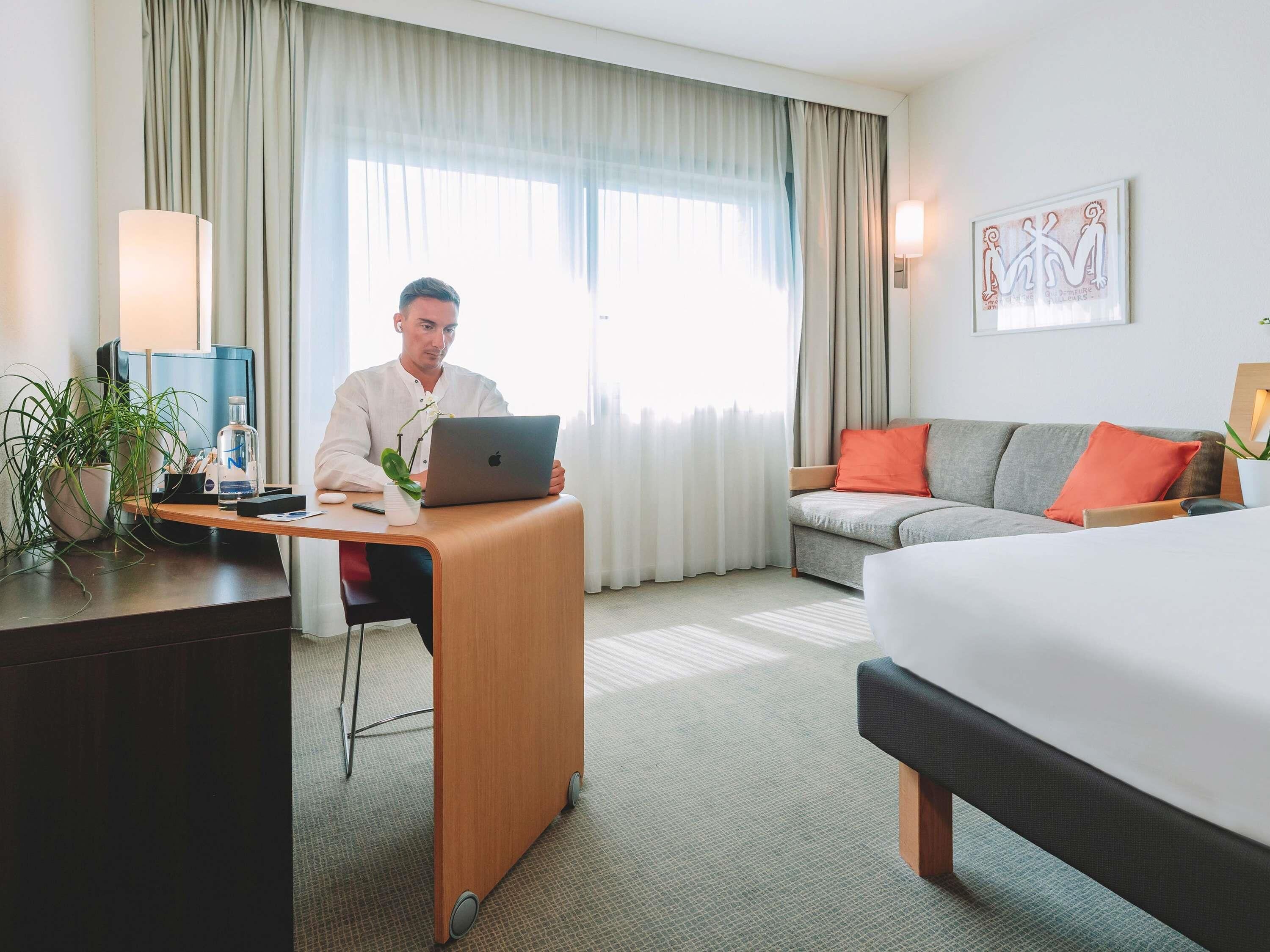 A hotel room with a desk, laptop, bed, sofa, and curtains.
