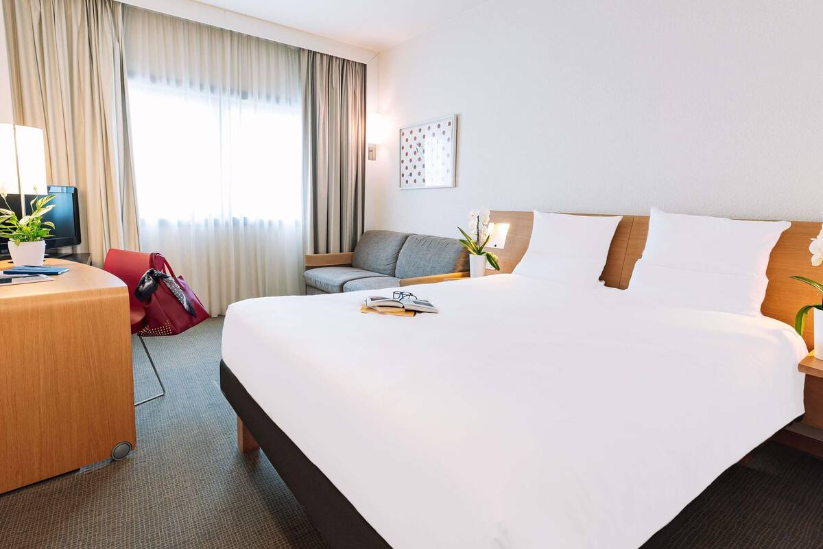 A hotel room with a large bed, a desk with a computer, a chair, and a potted plant.