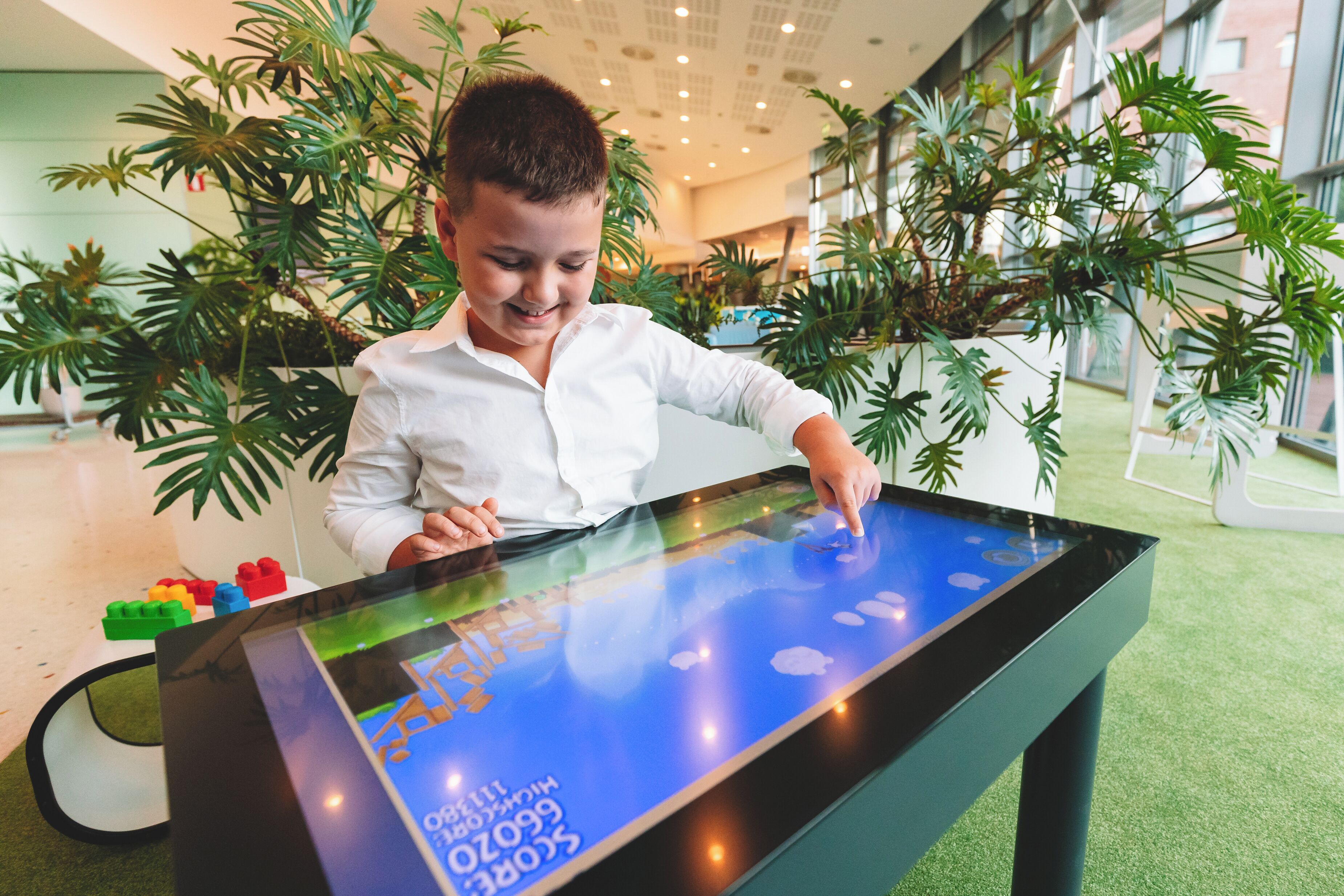 A child playing on a digital interactive table in a hotel lobby with indoor plants and a modern design.