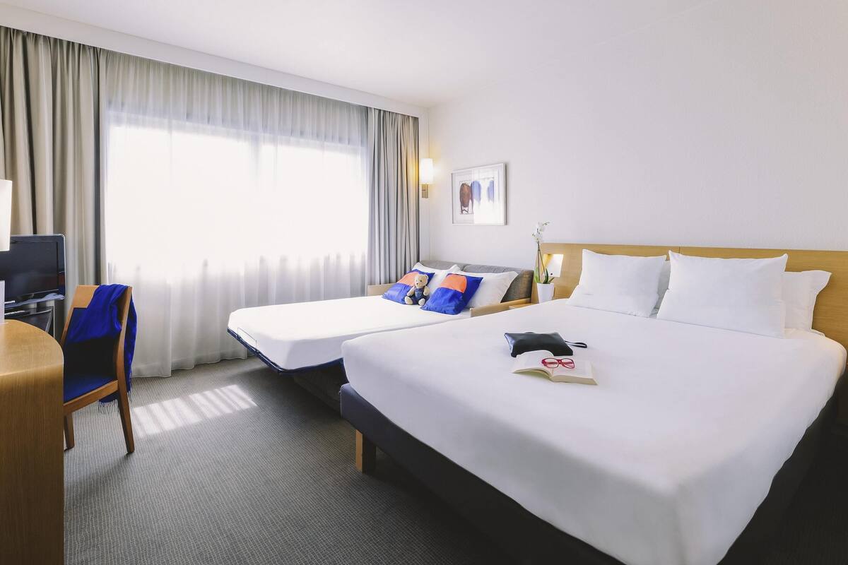 A hotel room with two beds, a desk, a chair, a TV, and a window with curtains.