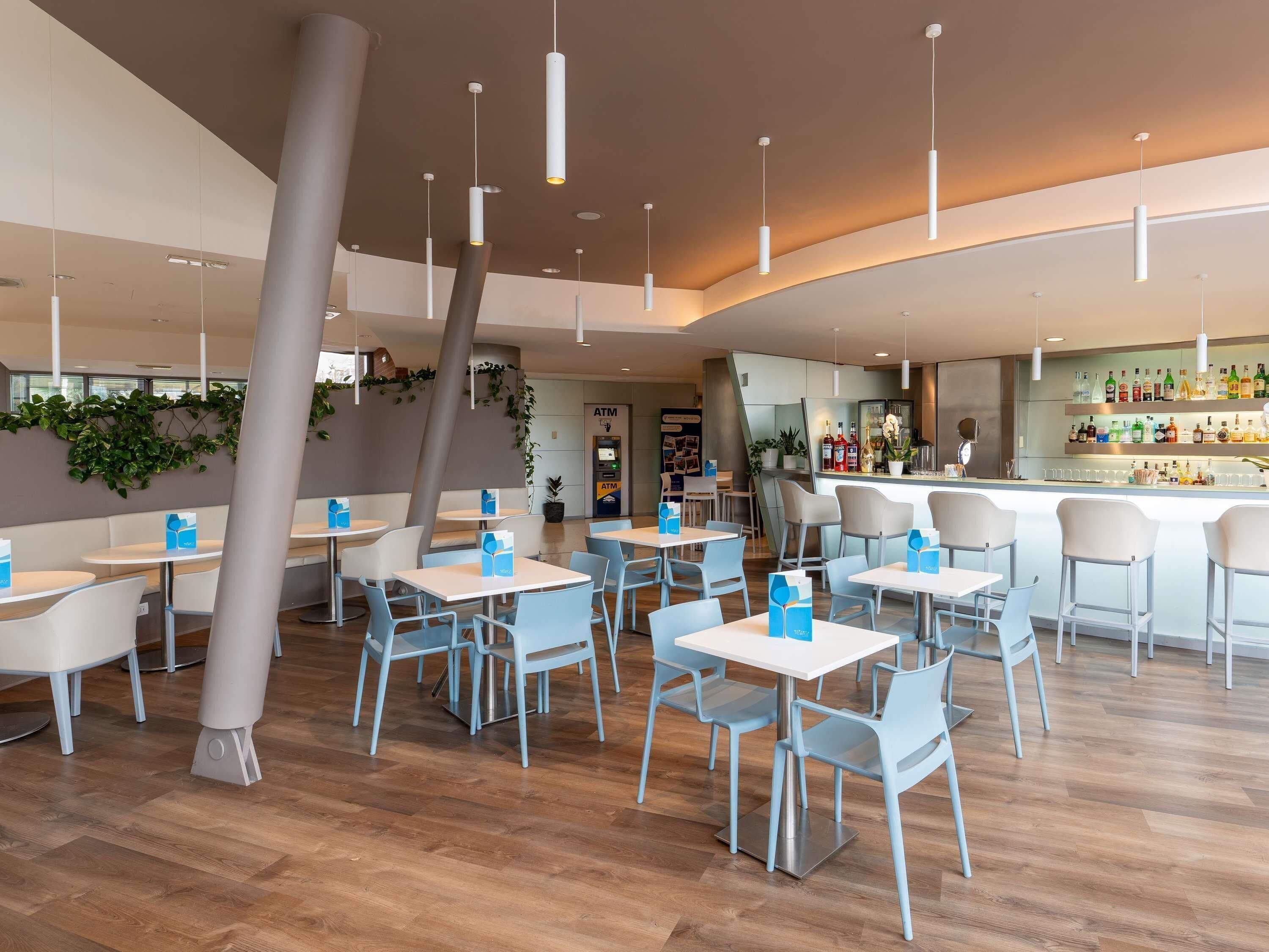 Interior moderno de restaurante con suelo de madera, sillas azul claro, mesas blancas, lámparas colgantes y una zona de bar con una variedad de botellas.