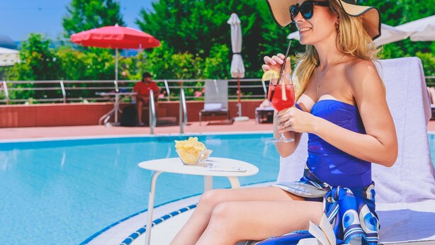 Una persona relajándose junto a una piscina con una bebida, un libro, un paraguas rojo y una pequeña mesa con patatas fritas.