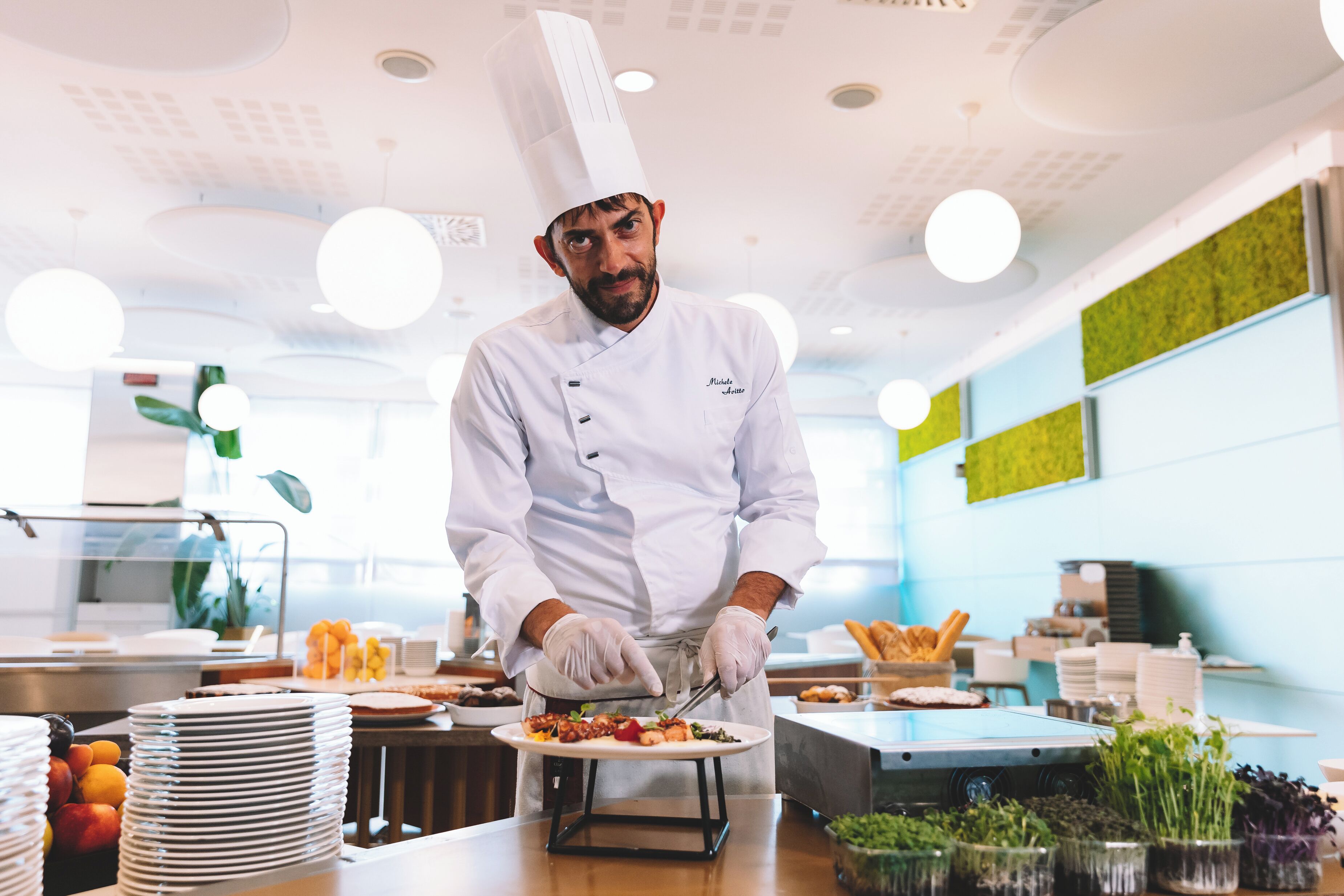 A chef in a white uniform and hat preparing food in a modern kitchen with a blue accent wall and various food items on the counter.