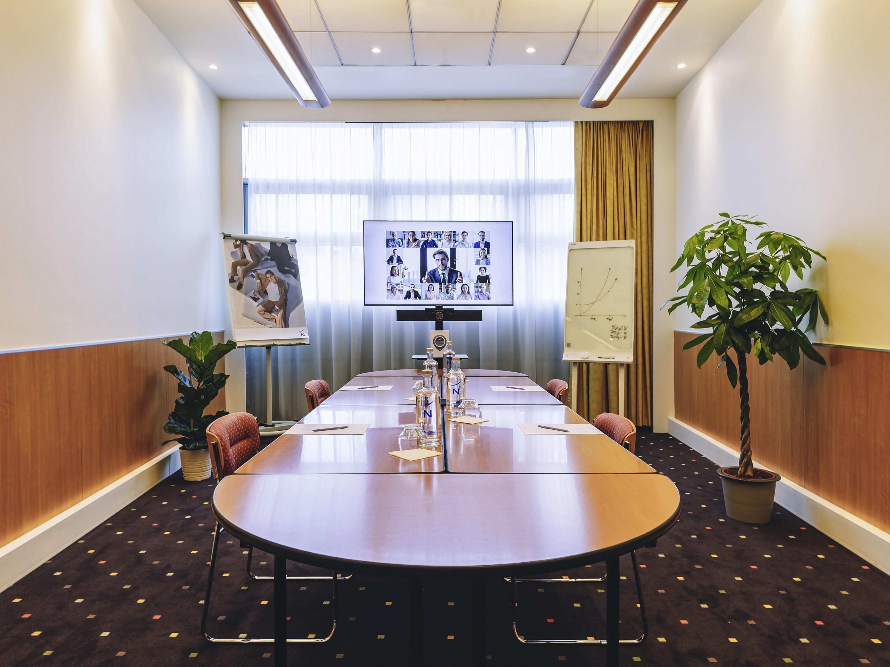 A conference room with a long table, chairs, a projector screen, a whiteboard, and a potted plant.