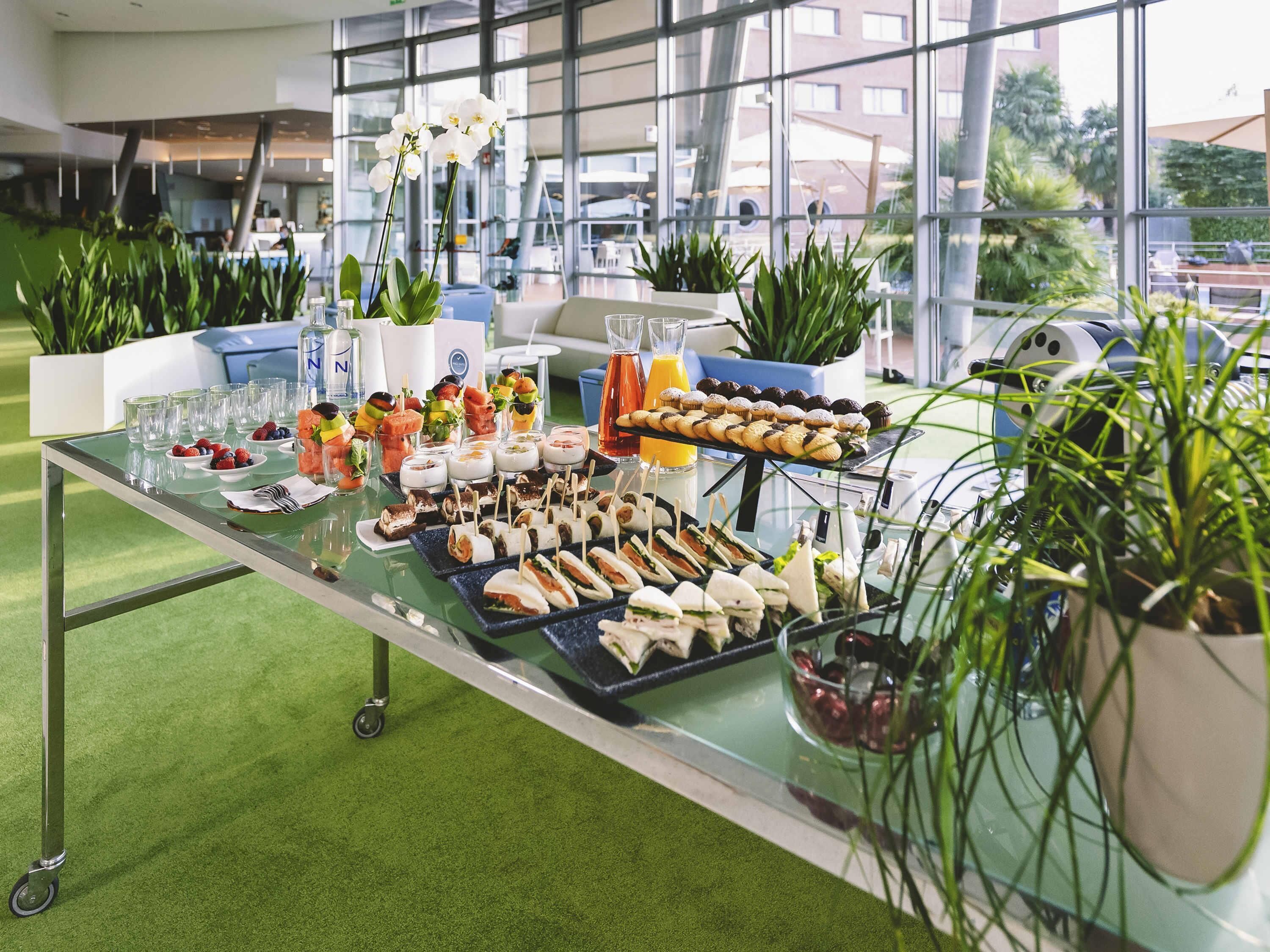 A buffet table with various food items and drinks, surrounded by potted plants and large windows.
