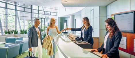 Una moderna zona de recepción de hotel con un mostrador de cristal, un área de asientos con sillas y un amplio ventanal que deja entrar luz natural.
