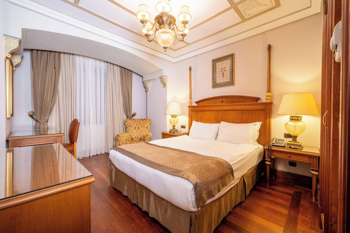 A hotel room with a large bed, a wooden desk, a chair, a nightstand with a lamp, and a chandelier.