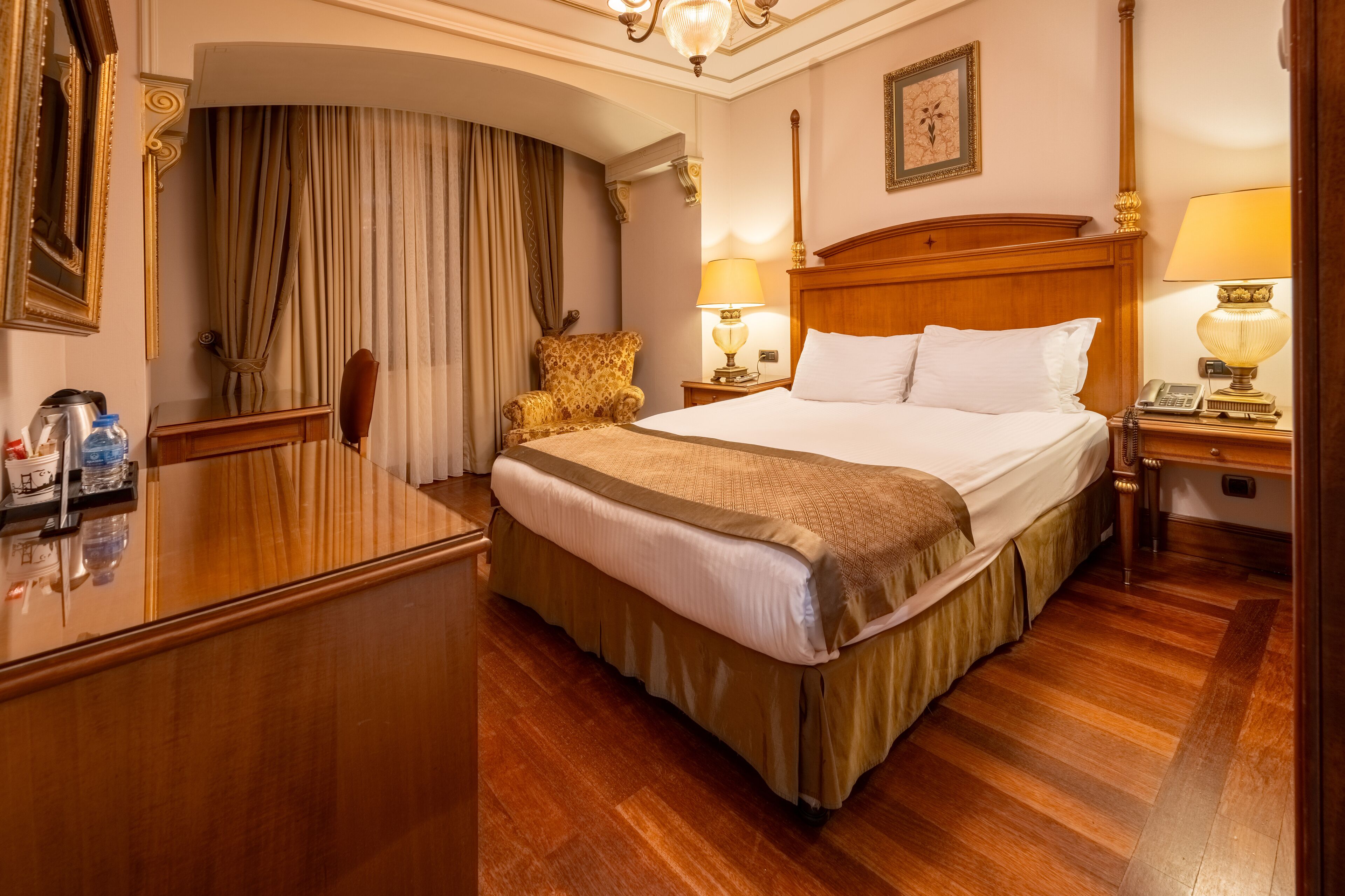 A hotel room with a large bed, wooden headboard, two bedside lamps, a wooden desk with a telephone, and a chair.