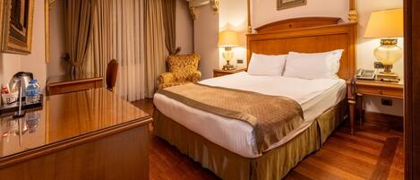 A hotel room with a large bed, wooden headboard, two bedside lamps, a wooden desk with a telephone, and a chair.