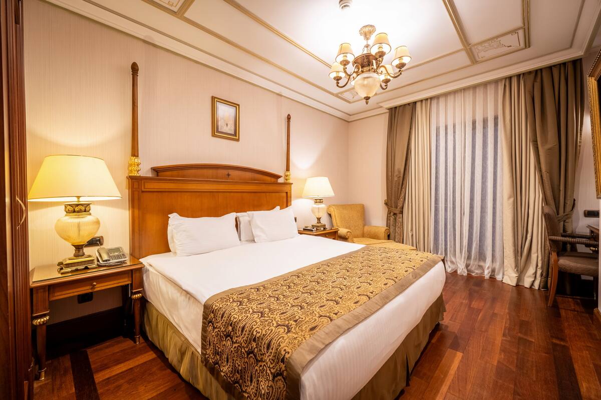 A hotel room with a large bed, two bedside tables with lamps, a wooden headboard, a chandelier, and a large window with curtains.