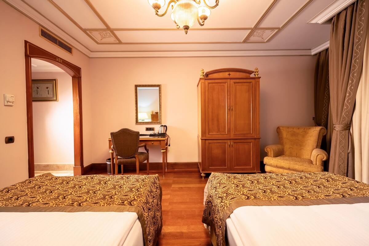 A hotel room with two beds, a desk, a chair, a wardrobe, and a chandelier.