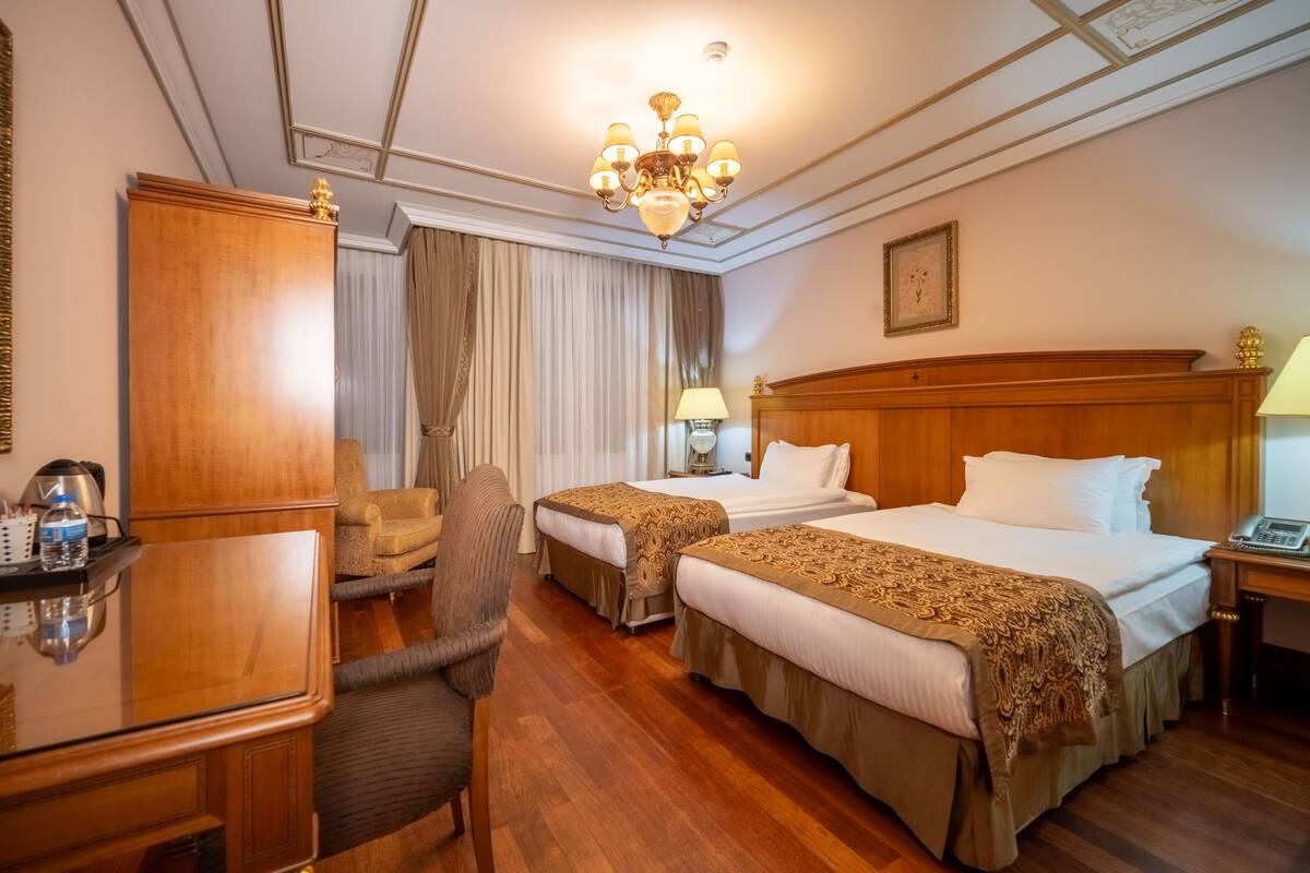 A hotel room with two beds, a desk, a chair, a chandelier, and a large window with curtains.
