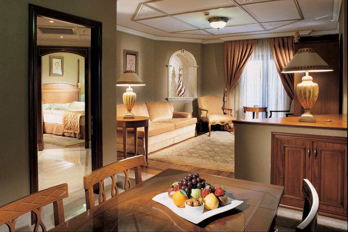 A hotel room with a bed, a sofa, a dining table with fruit, and a desk with a lamp.