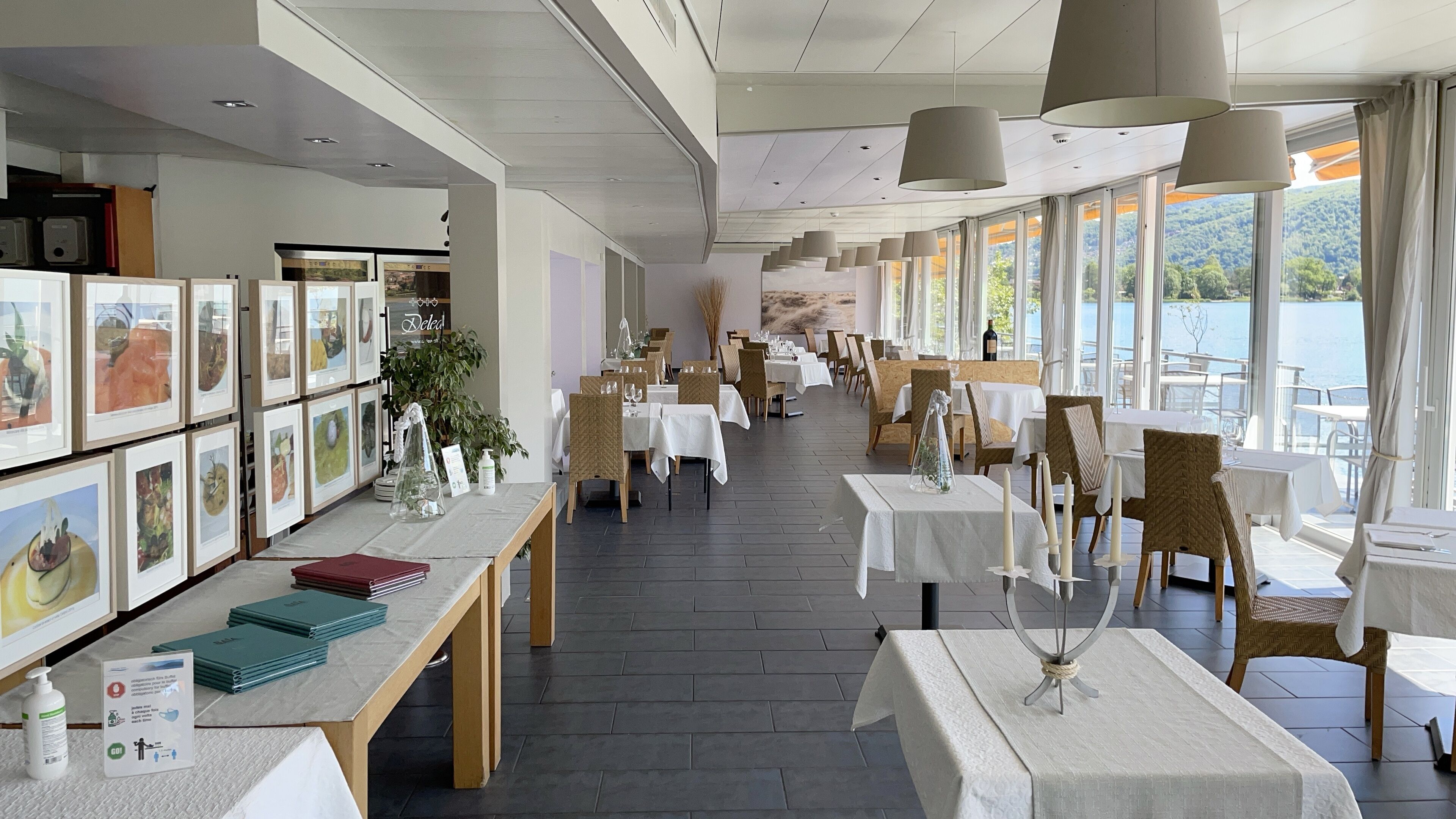 A restaurant with multiple tables and chairs, framed artwork on the walls, and large windows offering a view of the outdoors.