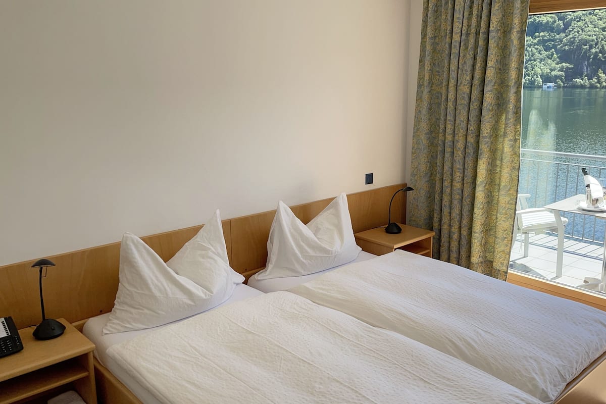 A hotel room with a bed, a nightstand, a lamp, and a view of a lake through a window.