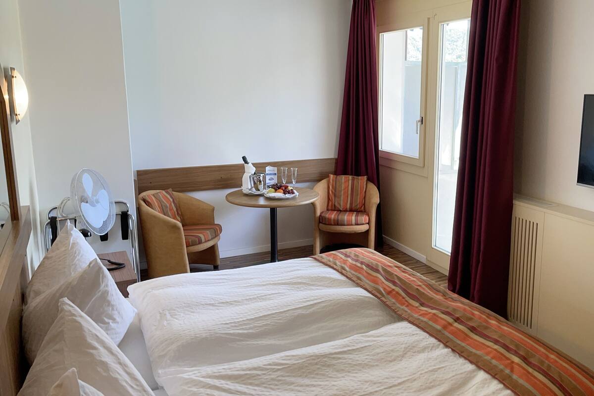 A hotel room with a bed, a small table with a bottle and glasses, two chairs, a TV, and a window with curtains.