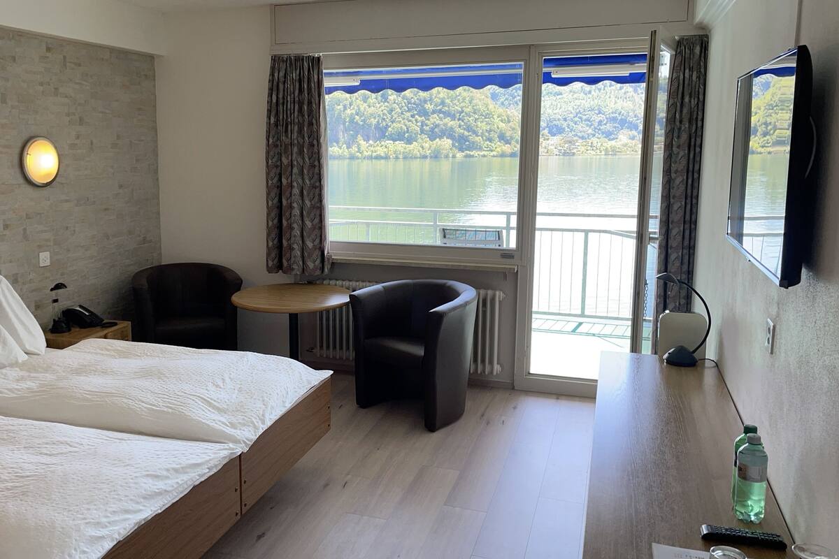A hotel room with a bed, a desk, a chair, a window with a view of a lake and hills, and a wall-mounted TV.