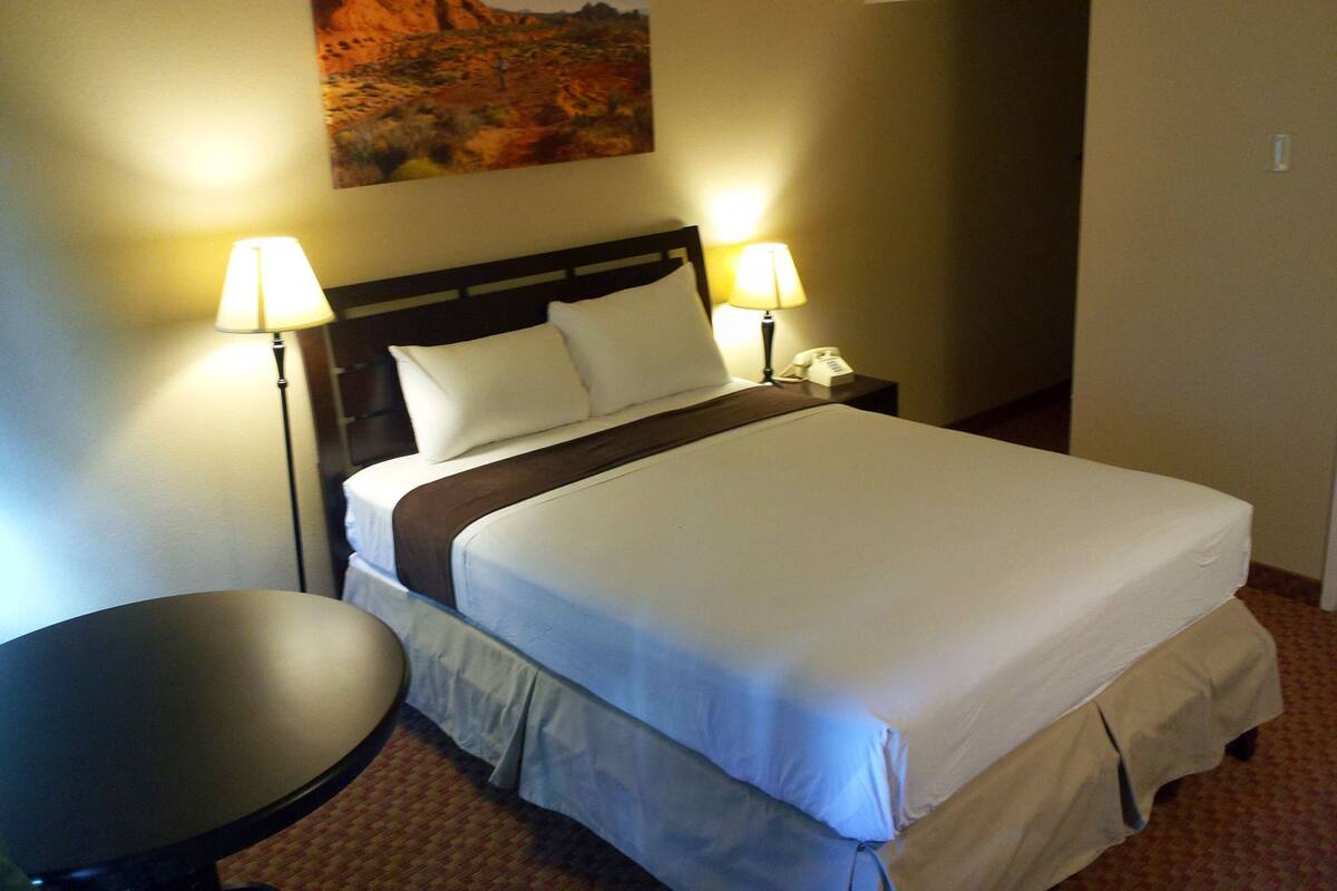 Uma cama bem arrumada com lençóis brancos, duas luminárias de cabeceira, uma mesinha redonda e um quadro de uma paisagem desértica na parede.